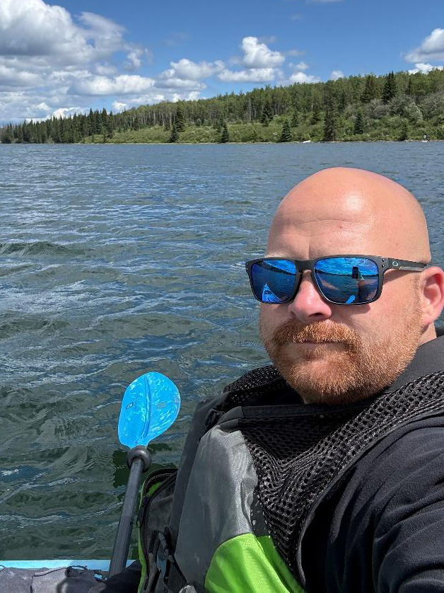 A man is out on a lake, looking at the camera while wearing sunglasses and a lifejacket and holding a kayak paddle