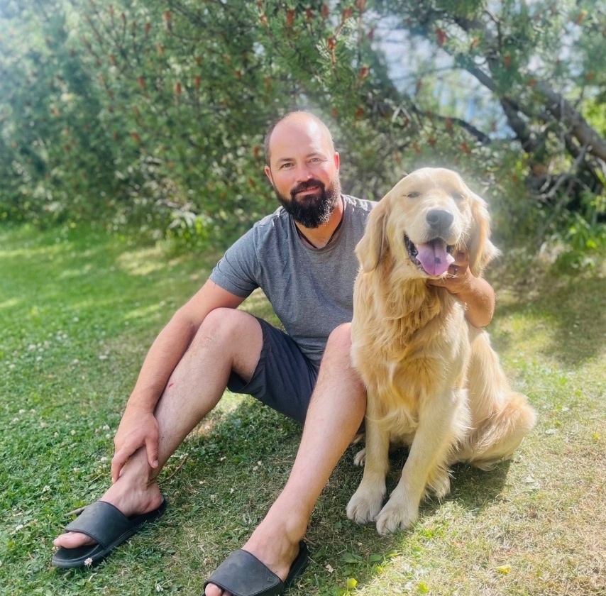 A man and his golden retriever are seated in the grass next to some bushes